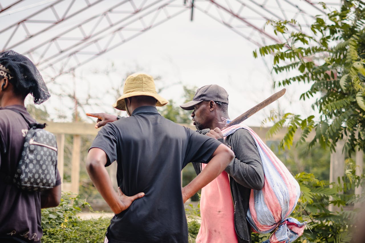 A group of three adults talking outdoors with greenery, one carrying a machete.